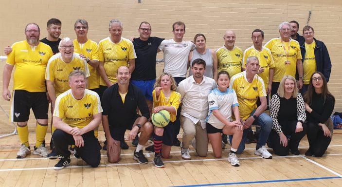 Group photo of a mixed team in yellow shirts and casual clothing, posing together in a gym.