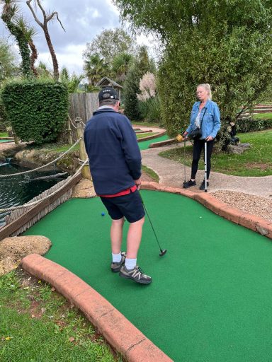 Two people playing mini golf on a green course with trees in the background.