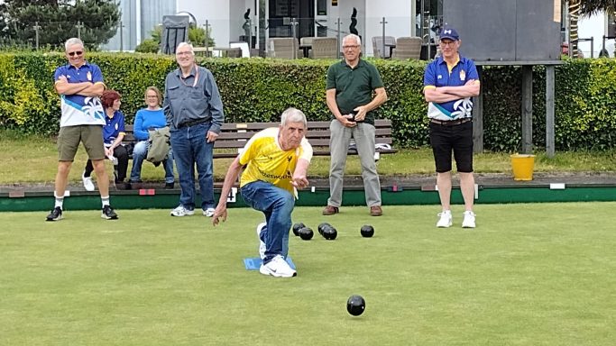 A player delivering a bowl on a green while spectators watch nearby.