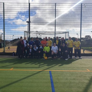 Group photo of a diverse football team on an artificial pitch with goalposts in the background.